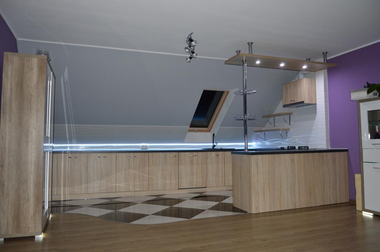 Jasna kuchnia na poddaszu z drewnianymi frontami, czarnym blatem i wyspą. Na podłodze wzór szachownicy. Nietypowe oświetlenie LED i okno dachowe.