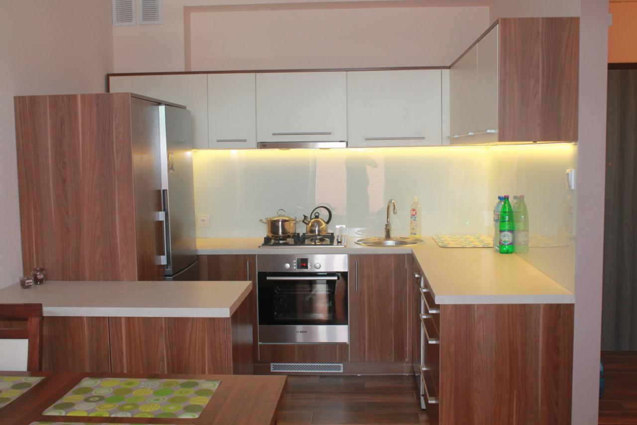 Nowoczesna kuchnia z drewnianymi frontami, białymi szafkami wiszącymi i podświetleniem LED. Na blacie czajnik i garnki na płycie grzewczej.