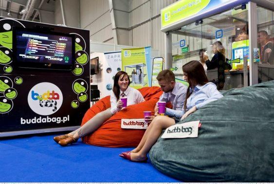Stoisko targowe z dużymi pufami do siedzenia marki Buddabag, na niebieskiej wykładzinie, z trzema osobami siedzącymi na pufach i trzymającymi fioletowe kubki, obok stanowiska z ekranem...