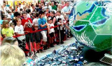 Dzieci otoczone czerwoną barierką obserwują dużą kulę ziemską zbudowaną z klocków Lego, na pierwszym planie rozsypane klocki Lego.