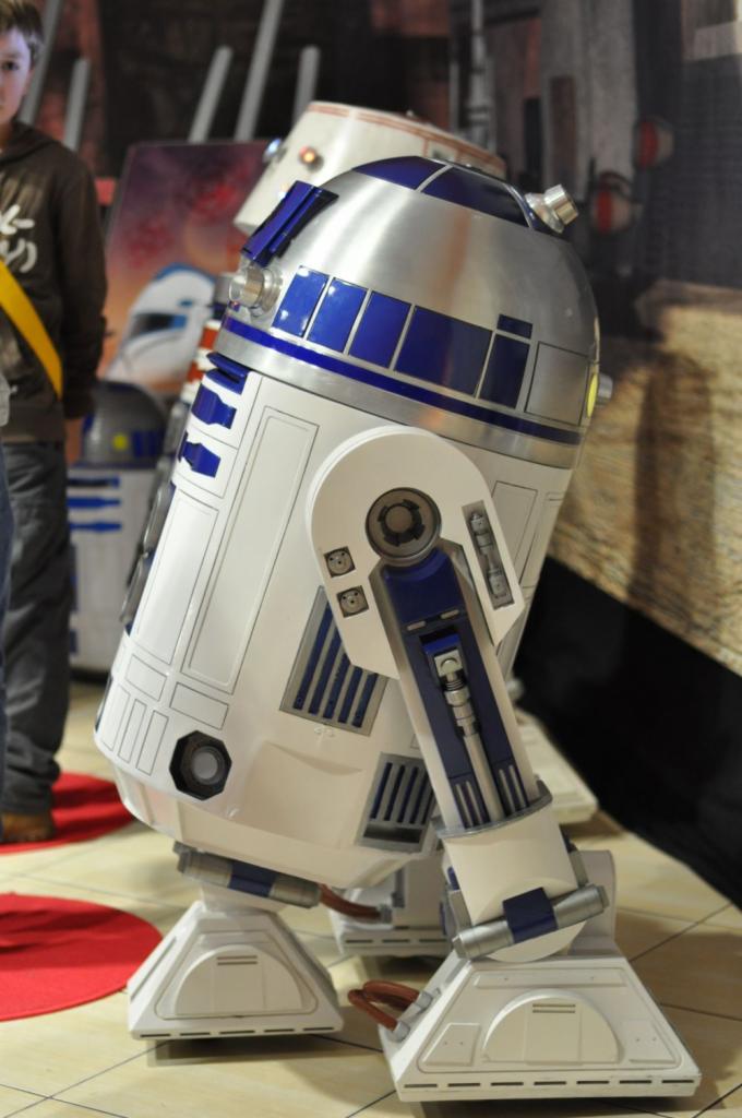 Szczegółowe ujęcie robota R2-D2 z filmu Star Wars na tle scenografii eventowej, widoczny fragment osoby w tle.