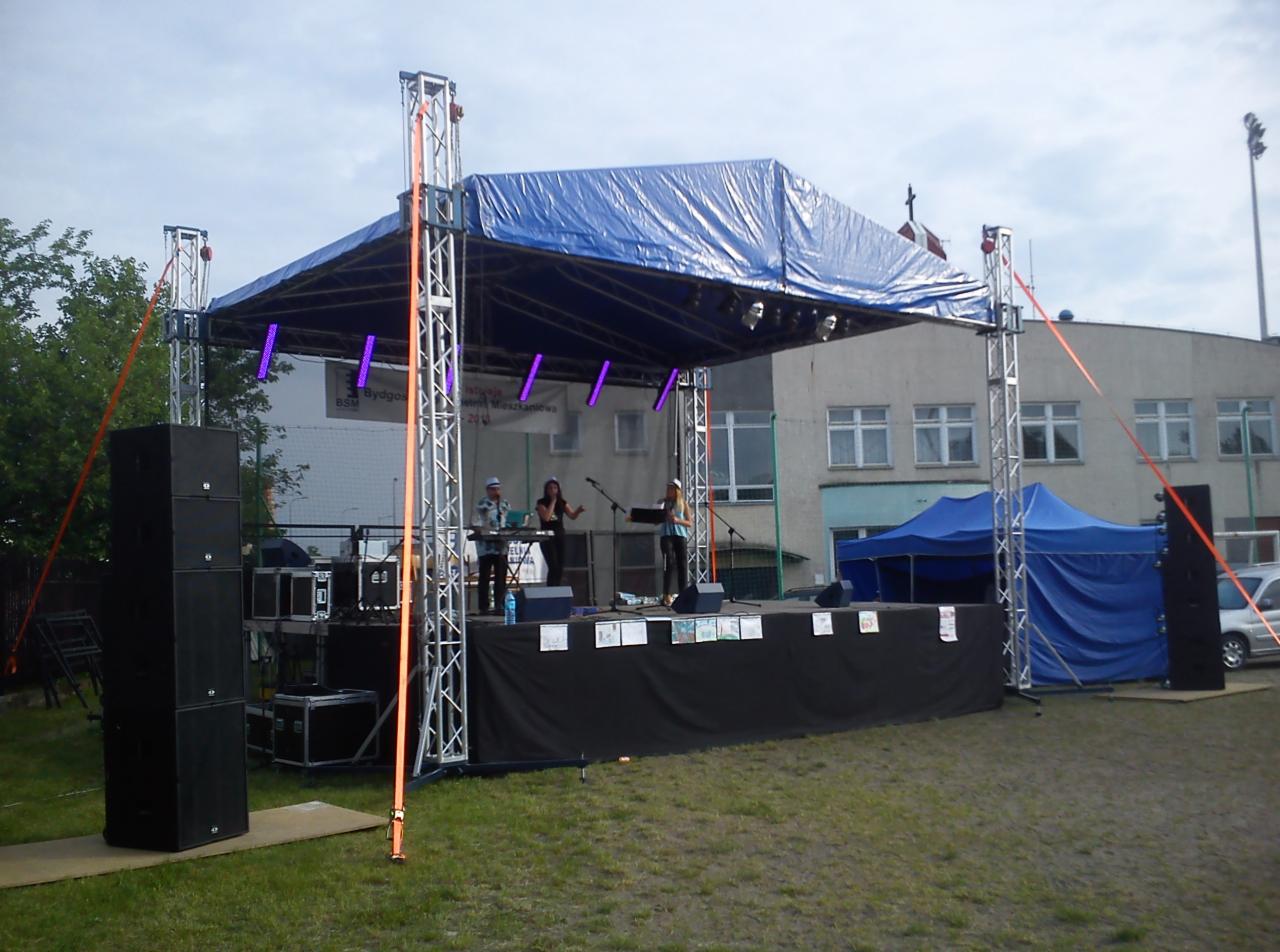 Zespół muzyczny na scenie z zadaszeniem z niebieskiej plandeki, nagłośnieniem i oświetleniem estradowym, na tle budynku z oknami i niebieskiego namiotu.