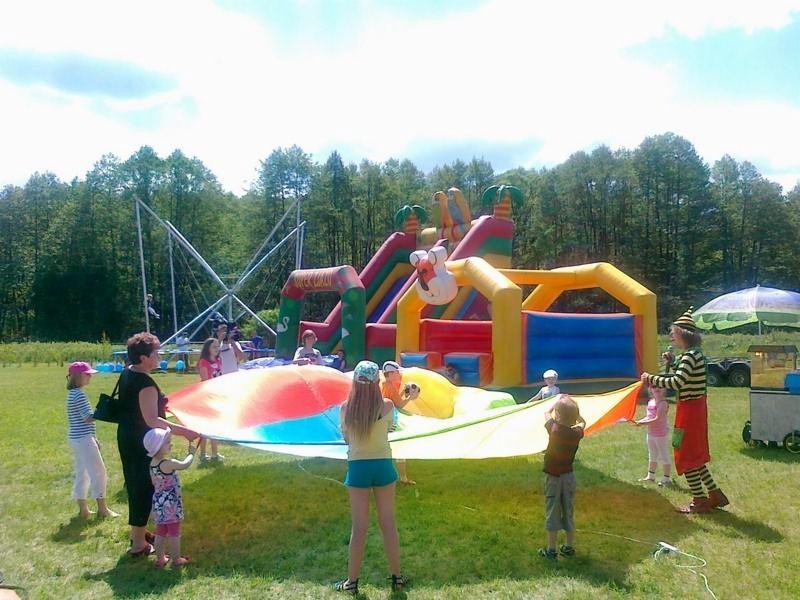 Animacje dla dzieci na zielonej trawie: grupa dzieci i dorosłych trzyma wielobarwną płachtę, w tle dmuchany zamek i trampolina.