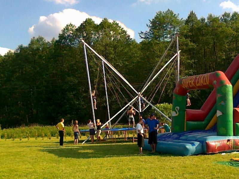 Rozrywka na świeżym powietrzu: trampolina bungee i dmuchana zjeżdżalnia na zielonej łące, widoczni ludzie w różnym wieku korzystający z atrakcji.