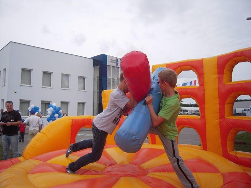 Dwóch chłopców walczy na dmuchanym ringu z użyciem dużych, miękkich pałek, otoczenie stanowi budynek biurowy i dekoracje z balonów.