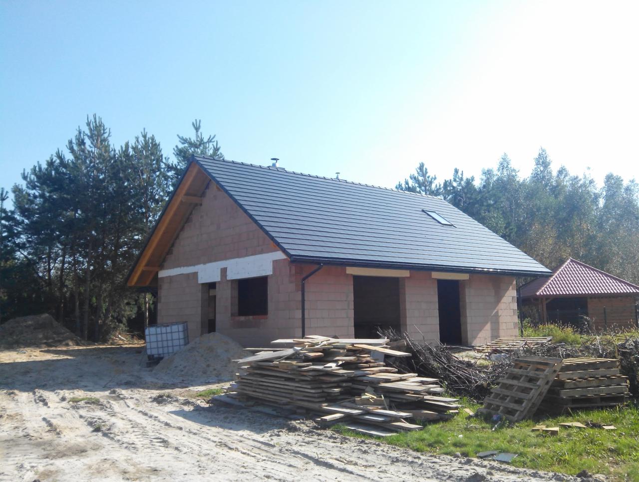 Surowy stan domu z bloczków z ciemną dachówką, widoczne drewniane elementy konstrukcji dachu, okna i drzwi niezamontowane, plac budowy z piaskiem i deskami w Głogowie Małopolskim.