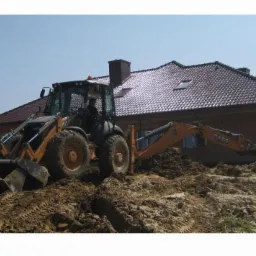 Pomarańczowa koparko-ładowarka Case na nierównym terenie z widocznymi śladami opon, w tle ceglany dom z ciemną dachówką i fragment lasu.