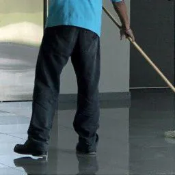 Pracownik w niebieskiej koszulce myje mopem błyszczącą, ciemną posadzkę w korytarzu biurowca, widoczne metalowe drzwi windy i gaśnica na ścianie.