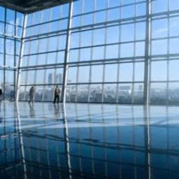 Przestronne, jasne wnętrze biurowe z polerowaną podłogą odbijającą ogromne okna i sylwetki trzech osób w oddali, widok na panoramę miasta za oknami.