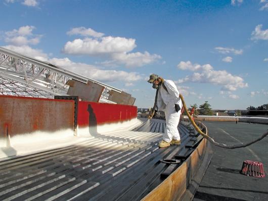Pracownik w kombinezonie ochronnym natryskuje pianę izolacyjną na metalową konstrukcję dachu, widoczne niebieskie niebo z chmurami i fragmenty konstrukcji stalowej.