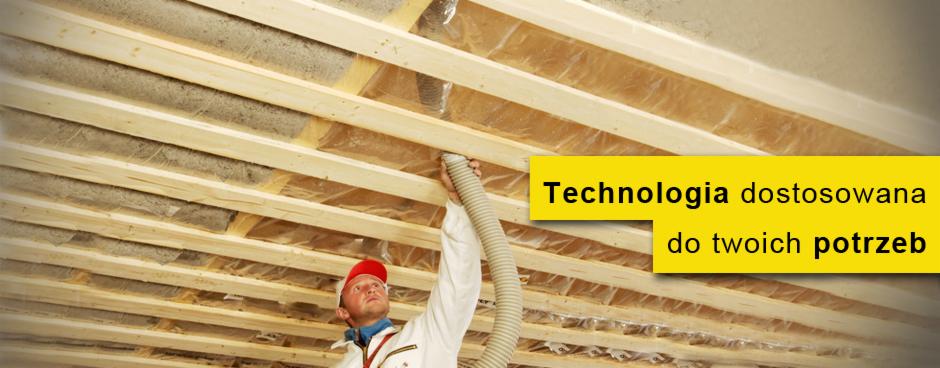 Pracownik w białym kombinezonie i czerwonej czapce aplikuje pianę izolacyjną w przestrzeni między krokwiami poddasza w Miękini, widoczne drewniane elementy konstrukcji dachu i folia paroizolacyjna.