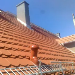 Czerwona dachówka karpiówka na skośnym dachu z kominem i wentylacją, widoczne rusztowanie zabezpieczające.