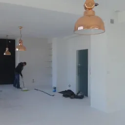 Mężczyzna maluje podłogę na biało w przestronnym, jasnym pomieszczeniu z trzema miedzianymi lampami wiszącymi, widoczne narzędzia malarskie i puszka z farbą.