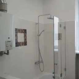 Biała łazienka z wanną i szklanym parawanem, widoczny piec gazowy Vaillant i dekoracyjne płytki z motywem kwiatowym.