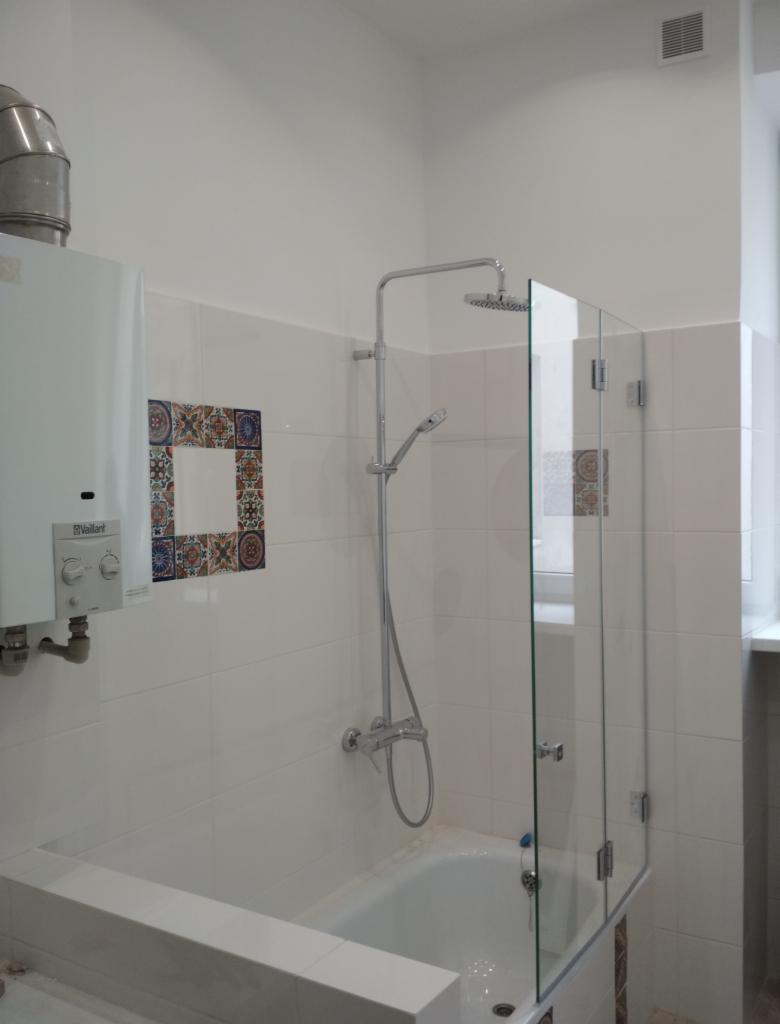 Biała łazienka z wanną i szklanym parawanem, widoczny piec gazowy Vaillant i dekoracyjne płytki z motywem kwiatowym.