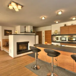 Jasne wnętrze domu z drewnianą podłogą, kominkiem obudowanym białym kamieniem, zabudową kuchenną z drewnianymi frontami i trzema hokerami przy wyspie.