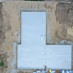 Widok z góry na płytę fundamentową w kształcie litery L, otoczoną drewnianym szalunkiem, z widocznymi elementami izolacji i instalacji, na placu budowy z ziemią i stertą drewna.