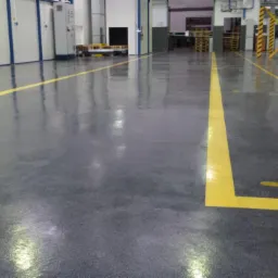 Błyszcząca, szara posadzka przemysłowa z żółtymi liniami wyznaczającymi strefy. Widoczne słupy z czarno-żółtymi pasami i palety w tle.