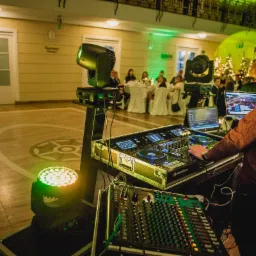 DJ obsługuje konsoletę podczas imprezy w sali balowej z oświetleniem scenicznym i gośćmi przy stołach.