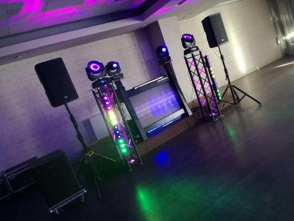 Sala z ustawionym sprzętem nagłośnieniowym i oświetleniowym, w tym kolumny głośnikowe na statywach, reflektory na kratownicach i konsoleta DJ-ska z ekranem.