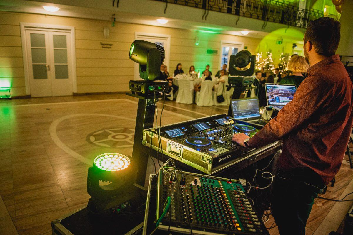 DJ obsługuje konsoletę podczas imprezy w sali balowej z oświetleniem scenicznym i gośćmi przy stołach.
