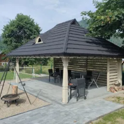 Kremowa altana z ciemnym dachem, wyposażona w stół i krzesła, grill oraz palenisko z trójnogiem na kamiennym podłożu. W tle budynek mieszkalny i drzewa.