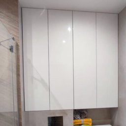 Firma ogólnobudowlana Grzegorz Groszek - Białe szafki wiszące nad pralką w łazience z kabiną prysznicową w tle, widoczny panel sterowania i ręczniki.