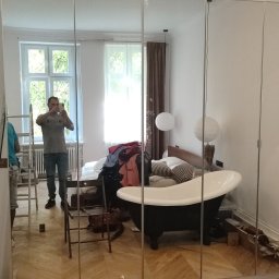 Firma ogólnobudowlana Grzegorz Groszek - Montaż lustrzanych drzwi przesuwnych szafy wnękowej, widoczne odbicie pokoju w trakcie remontu, drabina i osoba robiąca zdjęcie telefonem.