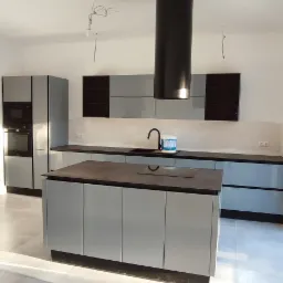 Nowoczesna kuchnia z wyspą i czarnym okapem cylindrycznym. Szare szafki z połyskiem, ciemny blat. Na blacie stoi wiadro z farbą. Białe ściany, szara podłoga.