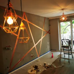 Ściana w trakcie malowania z geometrycznym wzorem w kolorze miedzianym, widoczne wiszące lampy w geometrycznych kloszach, zabezpieczona podłoga i akcesoria malarskie