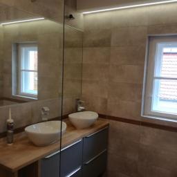 Firma ogólnobudowlana Grzegorz Groszek - Nowoczesna łazienka z dwoma umywalkami nablatowymi, szarą szafką pod umywalkę z drewnianym blatem, jasnymi płytkami na ścianach i oknem z widokiem na czerwony dach.