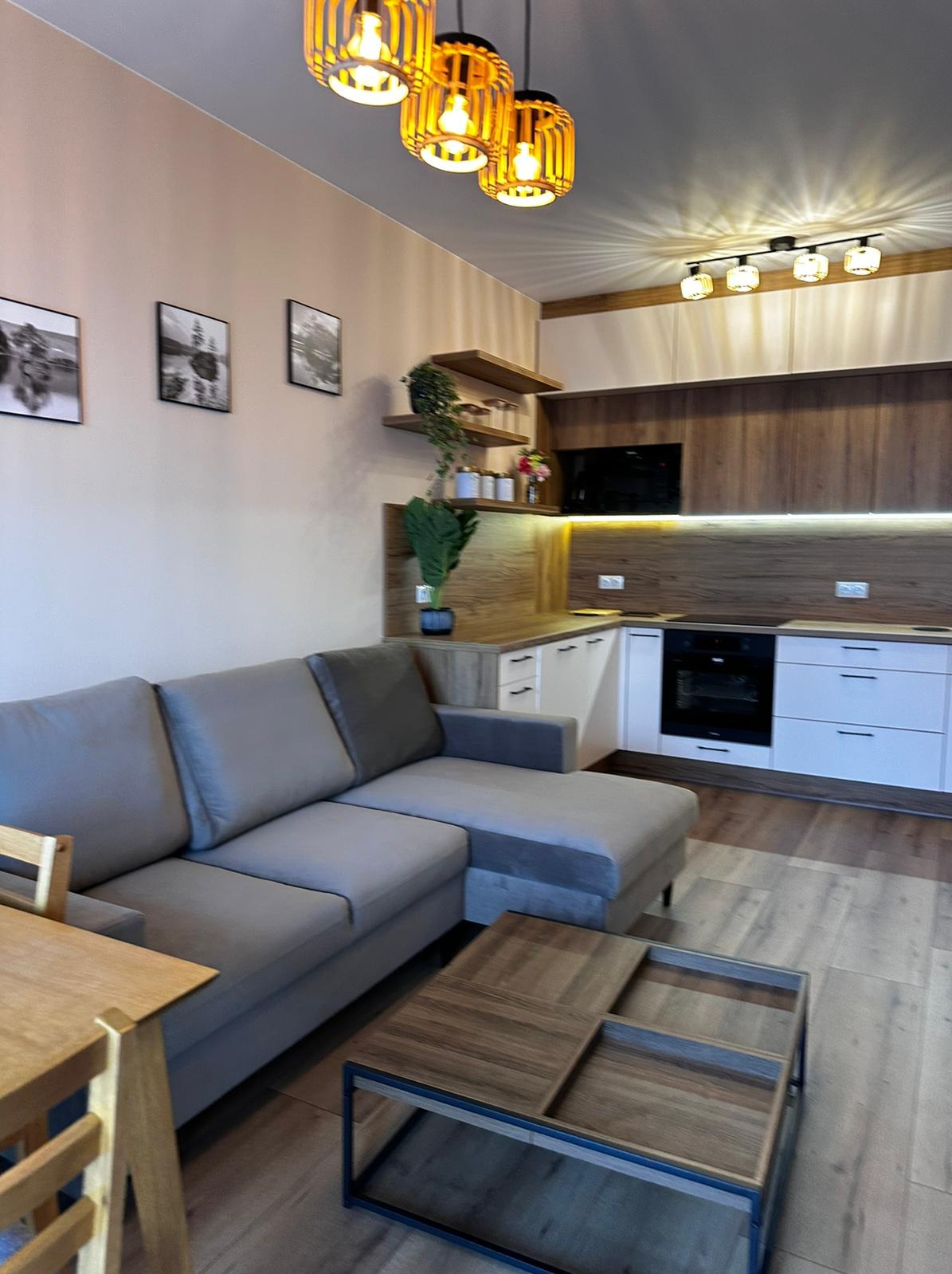 Salon z aneksem kuchennym: szara sofa narożna, drewniany stolik kawowy, zabudowa kuchenna z białymi frontami i drewnopodobnym blatem, oświetlenie punktowe i wiszące lampy z bambusa.