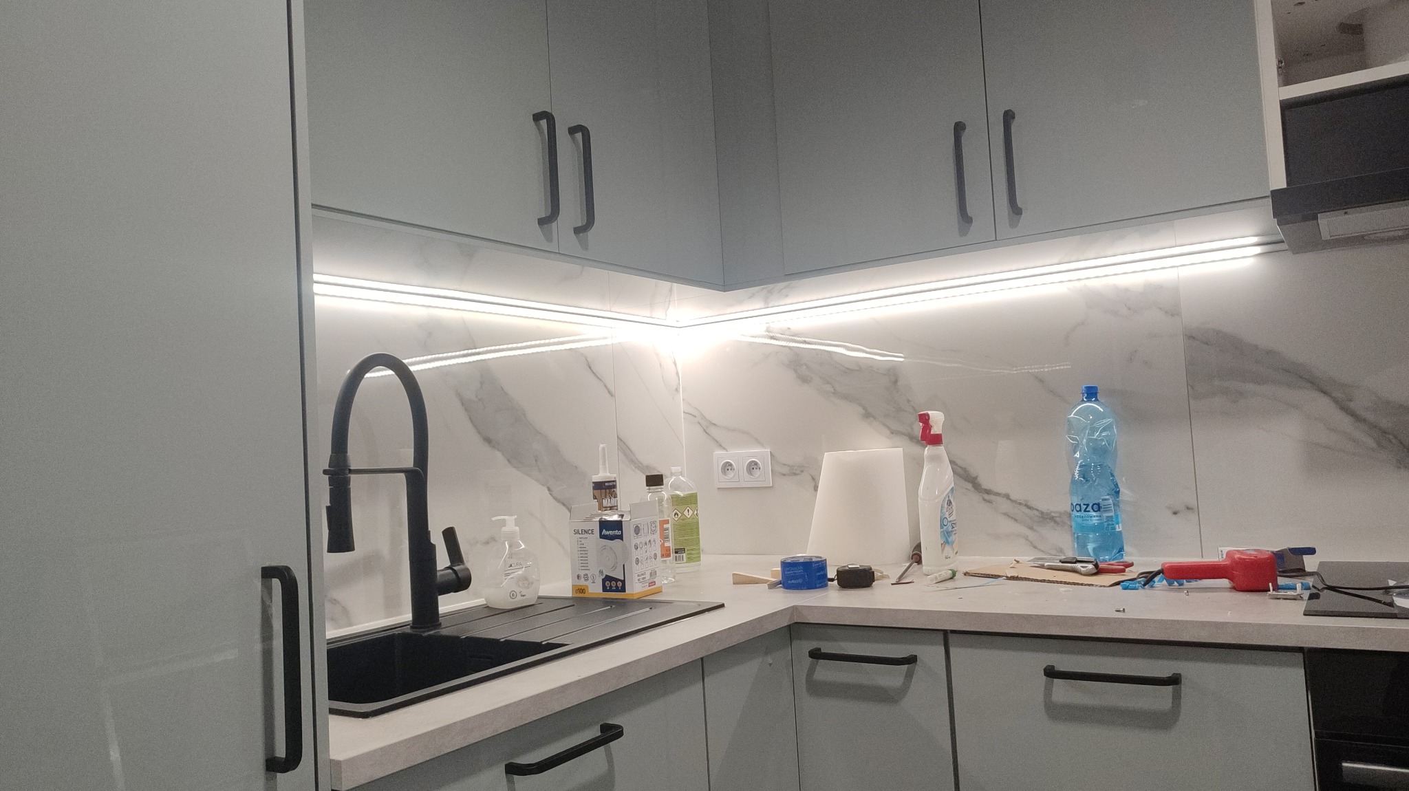Szare szafki kuchenne z czarnymi uchwytami i podświetleniem LED, marmurowa ściana, czarny zlew z baterią, widoczne akcesoria i narzędzia na blacie.