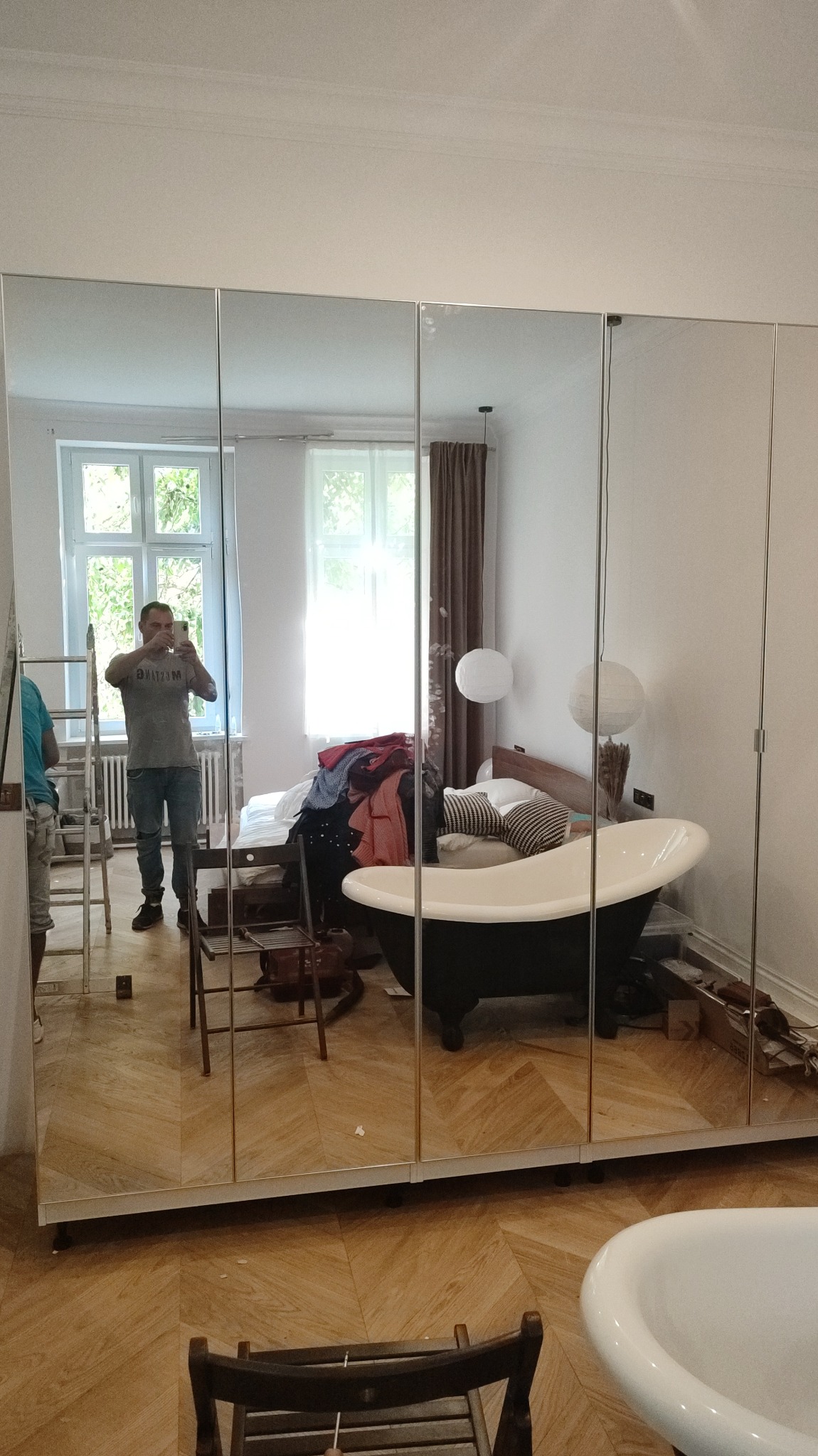 Montaż lustrzanych drzwi przesuwnych szafy wnękowej, widoczne odbicie pokoju w trakcie remontu, drabina i osoba robiąca zdjęcie telefonem.