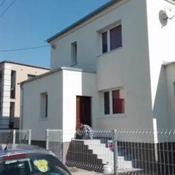 Dwukondygnacyjny dom z jasną elewacją, widok zza metalowego ogrodzenia, pracownik maluje schody wejściowe, w tle zaparkowany srebrny samochód osobowy.