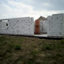 Wznoszone ściany z białych bloczków na fundamencie z czarną izolacją, fragment komina z czerwonej cegły, teren budowy z widoczną trawą i sąsiednim domem.