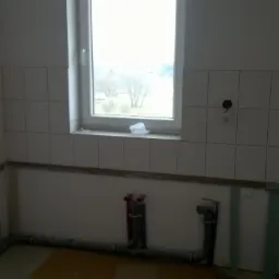 Wnętrze łazienki w trakcie remontu: białe płytki na ścianach, widoczne rury instalacji wodnej i przygotowane podłoże pod zabudowę. Okno z widokiem na zewnątrz.