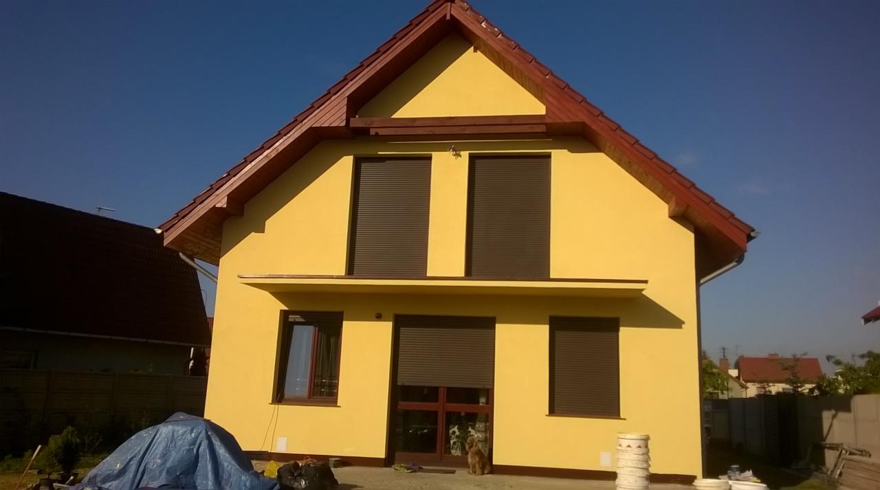 Żółty dom z brązowymi roletami i dachem z czerwonej dachówki w słoneczny dzień. Widoczne elementy elewacji i stolarki okiennej. Na podwórku leżą narzędzia.