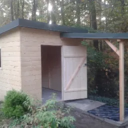 Surowa, drewniana altana z otwartymi drzwiami i zadaszeniem, na tle leśnego otoczenia. Widoczny fragment okna i betonowe podłoże. Naturalne światło słoneczne.