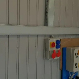 Panel sterowania bramą garażową Hormann A445 z łańcuchem zwisającym obok, skrzynka z gniazdami siłowymi (czerwone i niebieskie) oraz skrzynka sterownicza z przyciskami na metalowej ścianie garażu.