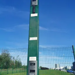 Zewnętrzny panel domofonowy z klawiaturą numeryczną i czytnikiem kart, zamontowany na zielonym słupku przy ogrodzeniu z siatki, na tle błękitnego nieba i zabudowań przemysłowych.