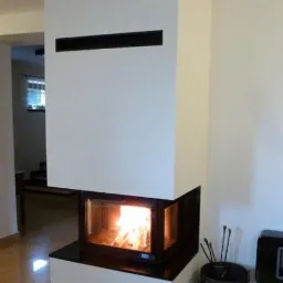 Nowoczesny, biały, narożny wkład z szybą, z czarnymi elementami wykończenia, z palącym się ogniem, wraz z akcesoriami kominkowymi w metalowym wiadrze.