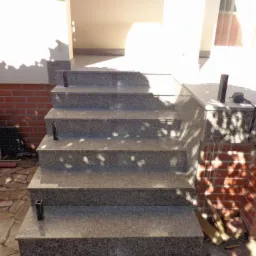 Granitowe schody zewnętrzne prowadzące do wejścia domu, wykończone płytkami z kamienia naturalnego, z widocznymi elementami metalowymi poręczy i cieniem liści na stopniach.