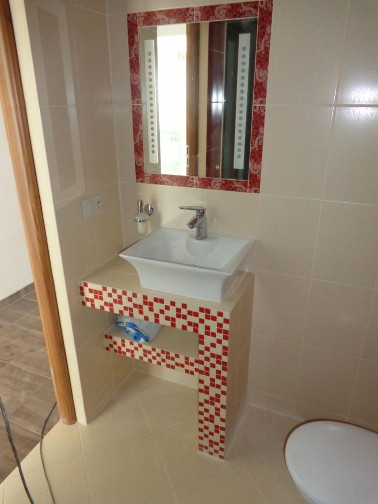 Umywalka nablatowa w kwadratowej szafce z czerwono-białą mozaiką, dopasowane lustro z oświetleniem LED w łazience z beżowymi płytkami.