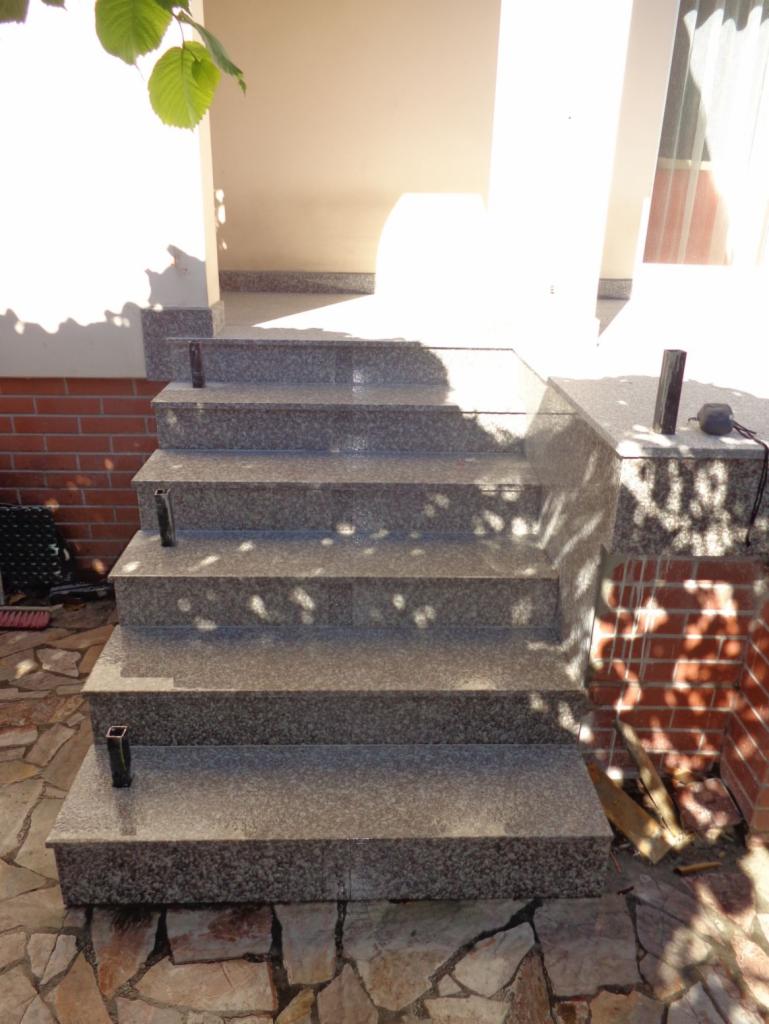 Granitowe schody zewnętrzne prowadzące do wejścia domu, wykończone płytkami z kamienia naturalnego, z widocznymi elementami metalowymi poręczy i cieniem liści na stopniach.