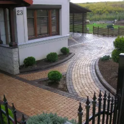 Aranżacja posesji z kostki brukowej w odcieniach beżu i brązu, z elementami małej architektury i zieleni. Widok na podjazd, chodnik i fragment ogrodzenia.
