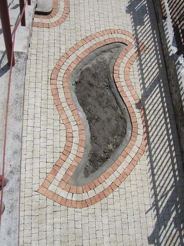 Kreatywnie ułożona kostka brukowa w odcieniach beżu i cegły, tworząca falisty wzór wokół przygotowanego miejsca na rośliny. Widok z góry, cień ogrodzenia.