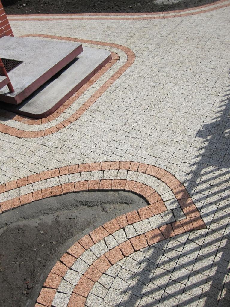 Jasna kostka brukowa ułożona w łukowate wzory z obrzeżami w odcieniach czerwieni, tworząca estetyczny chodnik z elementami małej architektury.