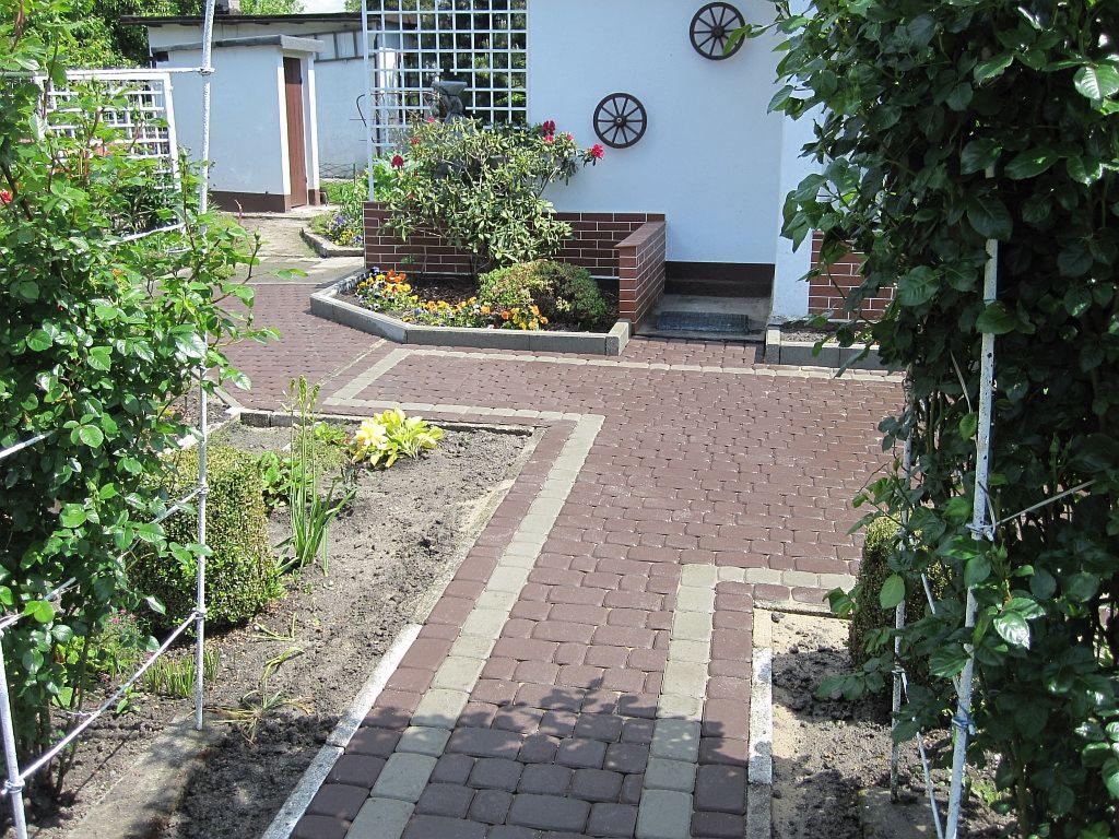 Czerwona kostka brukowa w ogrodzie, obrzeża z szarej kostki, rabaty kwiatowe, biała ściana budynku z dekoracjami. Ujęcie z poziomu gruntu, w słoneczny dzień.