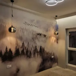 Pokój z fototapetą przedstawiającą mglisty las, oświetlony nowoczesną lampą sufitową z trzema okręgami LED i dwoma wiszącymi lampami z przydymionego szkła, surowa podłoga przed położeniem wykończenia.